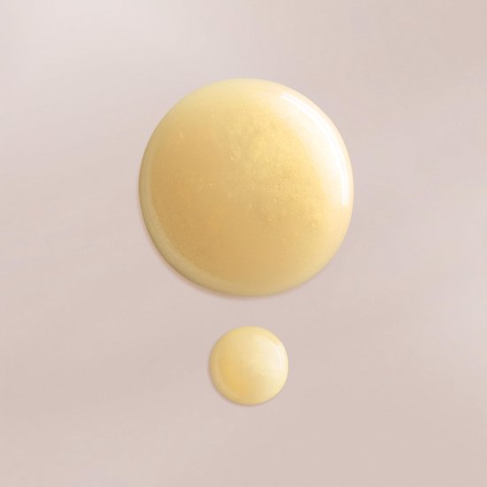 Twee druppels goudkleurig, glanzend serum op een lichtbeige achtergrond.