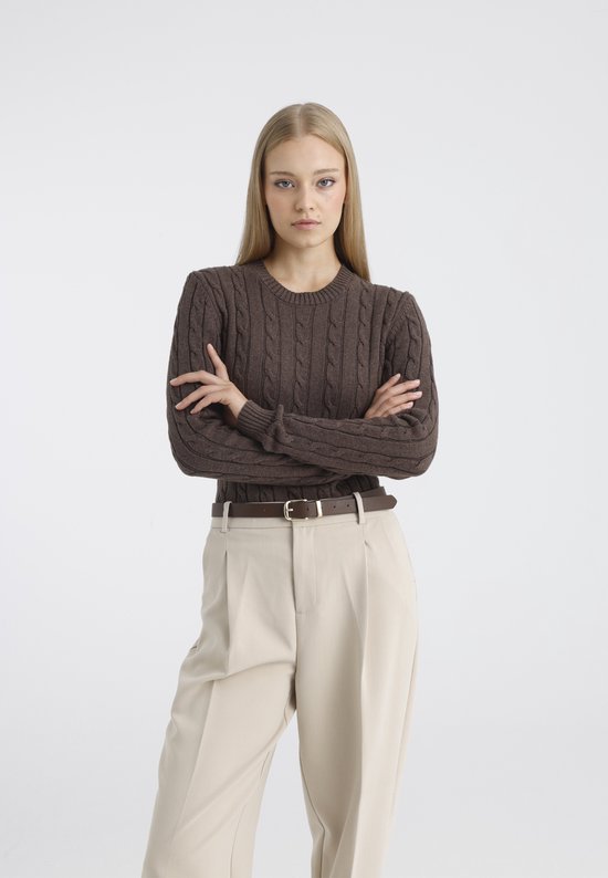 Model met bruine kabeltrui, beige broek en bruine riem, staand met armen over elkaar.