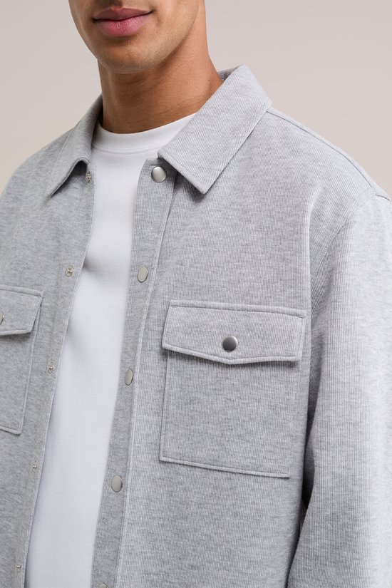 Close-up van een grijs fijngebreid overshirt met zilverkleurige drukknopen en opgestikte borstzakken.