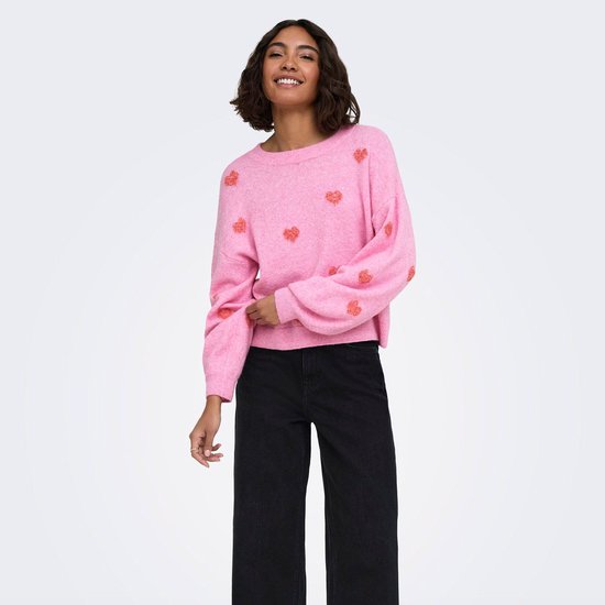 Een model draagt een roze trui met lange mouwen en rode hartjesprint. Ze draagt een zwarte broek.