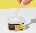 Een hand pakt met een houten spateltje wat witte crème uit een potje, de crème is draderig.