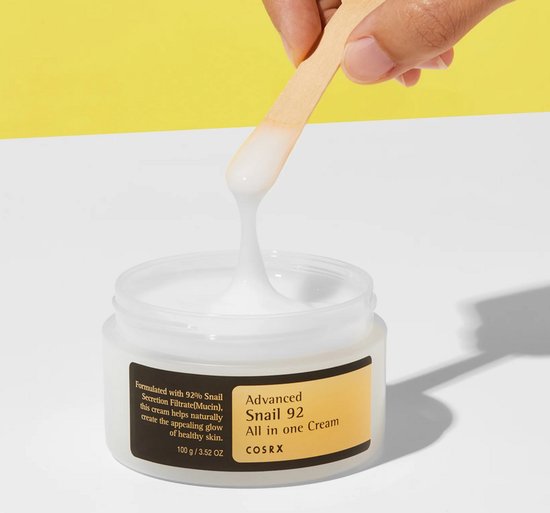 Een hand pakt met een houten spateltje wat witte crème uit een potje, de crème is draderig.