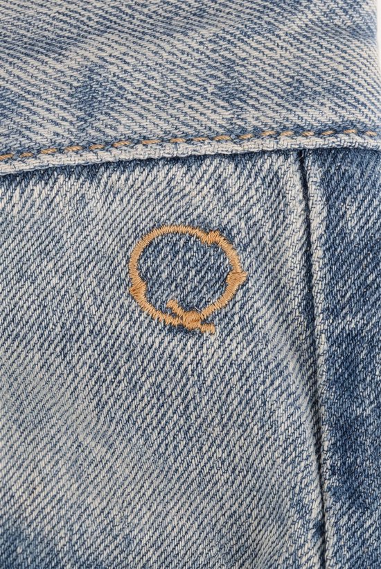 Detailopname van een lichtblauwe spijkerbroek met een goudkleurig, geborduurd cirkelvormig logo.