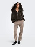 Model met lange krullen draagt een bruine nepbont jas met ritssluiting en een beige broek.