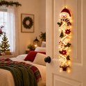 Kerstdeurhanger met sneeuwpop, bloemen en LED-verlichting opgehangen aan een witte deur in een slaapkamer.