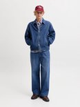 Man met korte blonde haren, donkerrode pet, jeansjack en wijde jeans, handen in de zakken.
