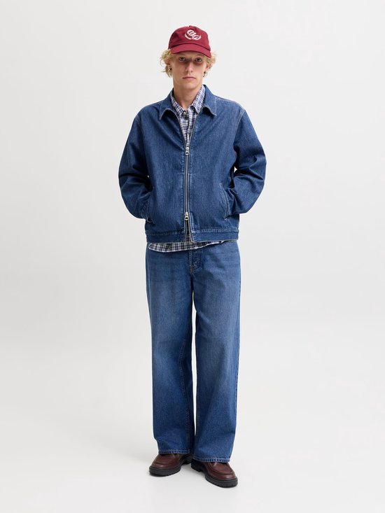 Man met korte blonde haren, donkerrode pet, jeansjack en wijde jeans, handen in de zakken.