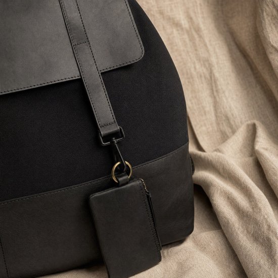 Detail van een zwarte rugzak met leren accenten en een stoffen etui aan een karabijnhaak.