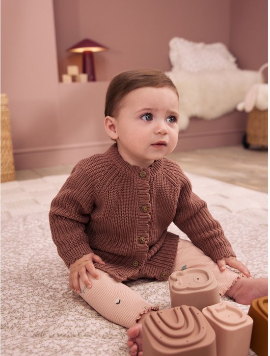 Baby in bruin gebreid vest met lichtroze broek en speelgoed.