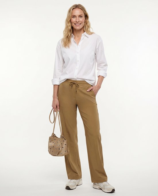 Model in wit overhemd en bruine travelstof broek met rechte pijpen, handtas in de hand.