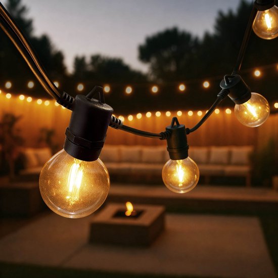 Een buitenlichtsnoer met 75 G40-lampen met E12-fitting, die warm wit licht uitstralen. De touwen hangen over een tuin met een vuurplaats en loungeset.