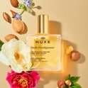 Nuxe Huile Prodigieuse multi-purpose dry oil in een glazen fles met gouden dop, omringd door bloemen en noten.