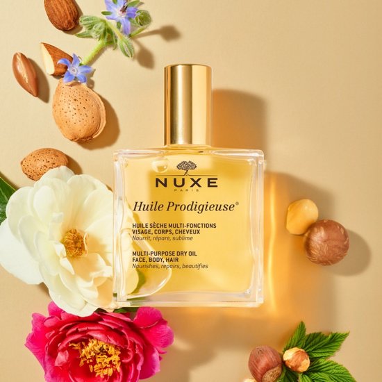 Nuxe Huile Prodigieuse multi-purpose dry oil in een glazen fles met gouden dop, omringd door bloemen en noten.