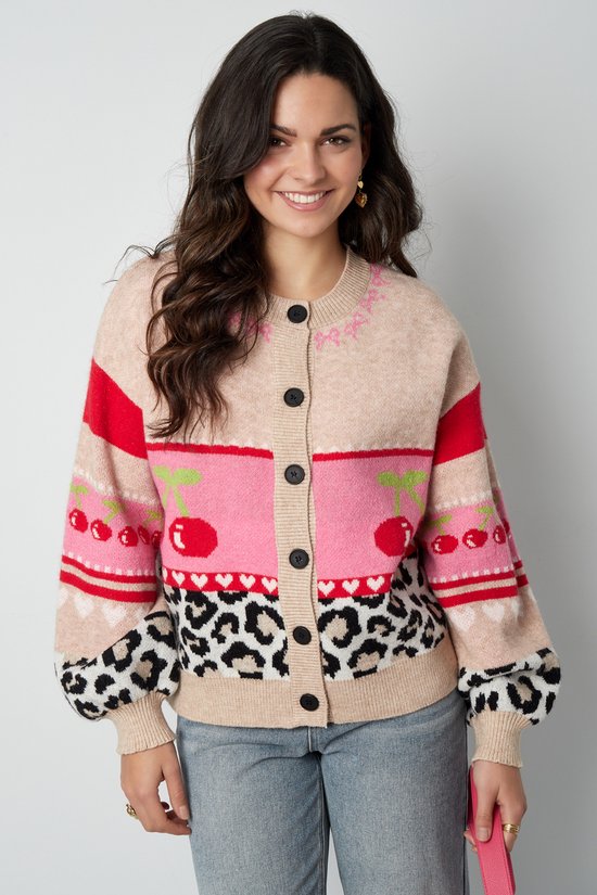 Model draagt beige cardigan met knopen. Ronde hals en pofmouwen. Kersen, hartjes en leopardprint op de body en mouwen.