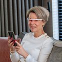 Vrouw met kort grijs haar draagt een AYO-bril met rood licht en houdt een smartphone vast.