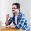 Man met blauw-geruite blouse, witte t-shirt, en een lichttherapiebril met rode accenten, drinkt koffie uit een mok.
