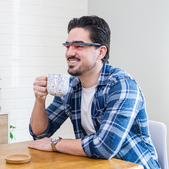 Man met blauw-geruite blouse, witte t-shirt, en een lichttherapiebril met rode accenten, drinkt koffie uit een mok.