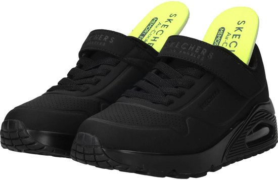 Paar zwarte Skechers sneakers met klittenbandsluiting en neongele binnenzolen met tekst.