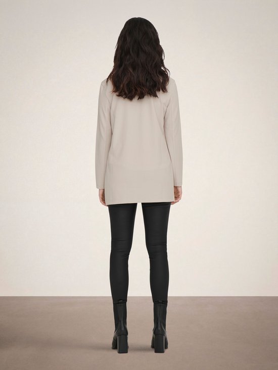 Vrouw van achteren gezien, met lange donkere haren, staand in een lange, beige blazer en zwarte broek.