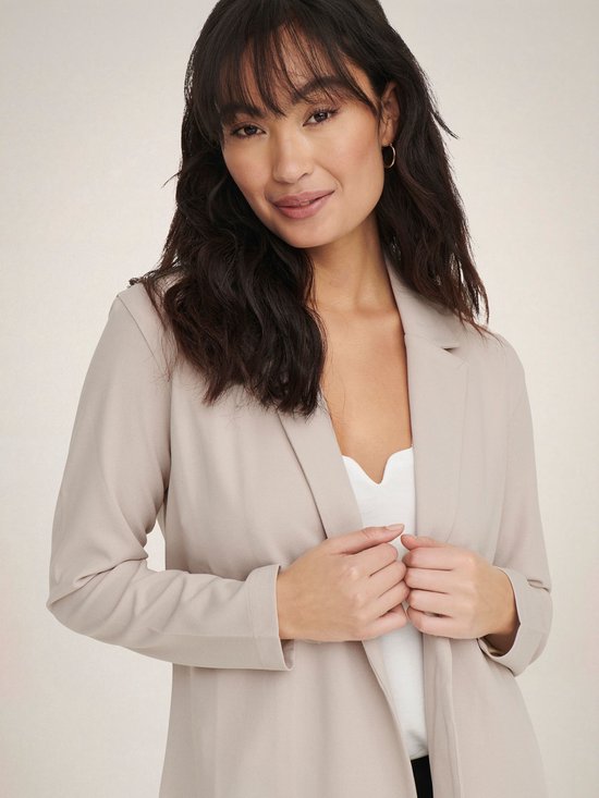 Vrouw met lange, golvende donkere haren in een lange, beige blazer met open voorkant en een witte top.