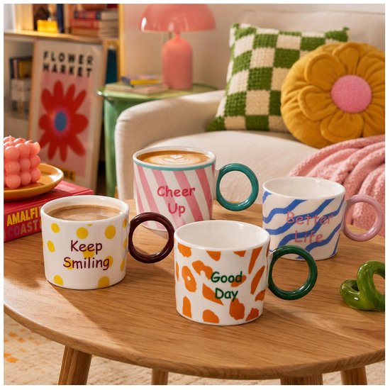 Vier mokken op een houten tafel. Ze hebben cirkelvormige handvatten en patronen: gele stippen met 'Keep Smiling', roze strepen met 'Cheer Up', oranje vlekken met 'Good Day' en blauwe golven met 'Better Life'.
