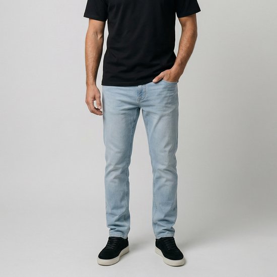 Man met de hand in de zak, staand in een lichtblauwe jeans en zwarte sneakers.