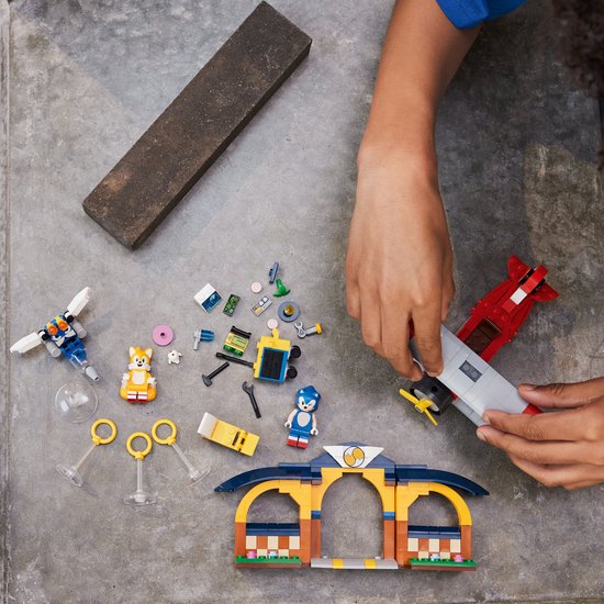 Handen bouwen de LEGO Sonic Tails' werkplaats en het Tornado-vliegtuig op een grijze tafel.