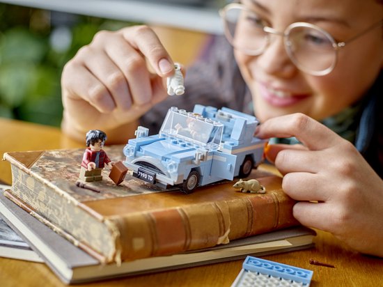 Une personne jeune portant des lunettes joue avec une Ford Anglia Lego bleu clair. Harry Potter, Ron Weasley, Hedwige et Croûtard sont des figurines, sur un livre.