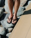 Bruine leren Reef slippers met zwarte zolen, gedragen door een man op rotsen bij een surfplank.