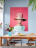Audrey Hepburn portant un grand chapeau de paille avec un nœud rose, une robe bleu pâle épaules dénudées, sur fond rose, au-dessus d'une table en bois.