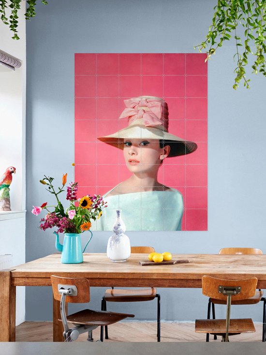 Audrey Hepburn portant un grand chapeau de paille avec un nœud rose, une robe bleu pâle épaules dénudées, sur fond rose, au-dessus d'une table en bois.
