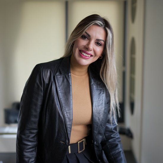 Vrouw met blond haar, draagt een zwarte leren jas en een camel coltrui.
