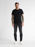 Man draagt een strakke zwarte spijkerbroek, zwarte T-shirt en witte sneakers. Volledige voorkant.