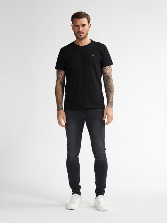 Man draagt een strakke zwarte spijkerbroek, zwarte T-shirt en witte sneakers. Volledige voorkant.