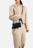 Zwarte portemonnee met klep, gedragen over de schouder door een model met beige jasje en wijde linnen broek.