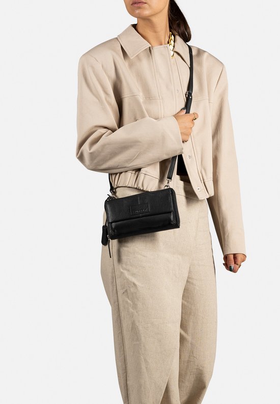 Zwarte portemonnee met klep, gedragen over de schouder door een model met beige jasje en wijde linnen broek.