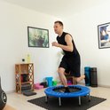 Een man in een zwart sportshirt en korte broek rent op een blauwe trampoline in een woonkamer.