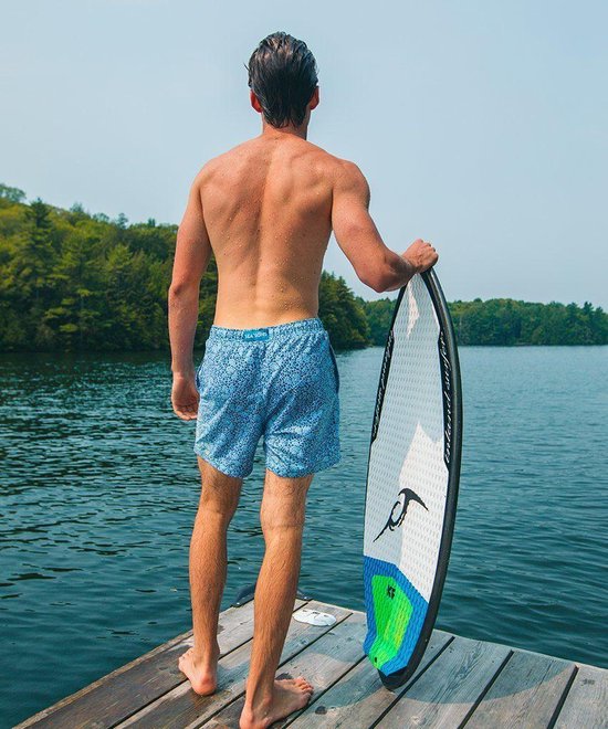 Man op een steiger met een surfplank, draagt een blauwe zwembroek met een all-over mozaïekprint.