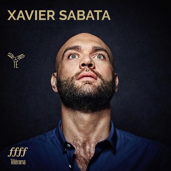 Close-up van Xavier Sabata, een kale man met baard, die omhoog kijkt. Hij draagt een marineblauw overhemd. "XAVIER SABATA" bovenaan.
