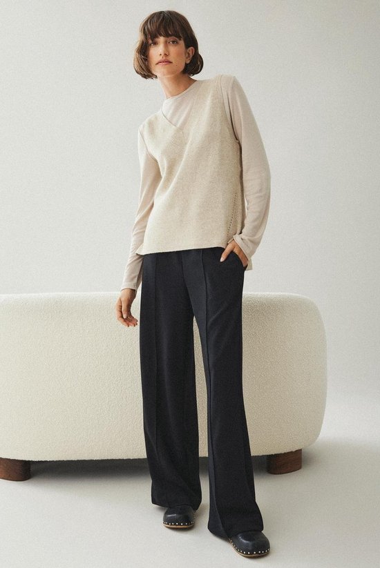 Model met beige gebreide trui zonder mouwen over een lichtbeige top met lange mouwen en een zwarte broek met wijde pijpen.
