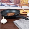 Zwarte koekenpan met granieten look met biefstuk, met knoflook en pepers ernaast.