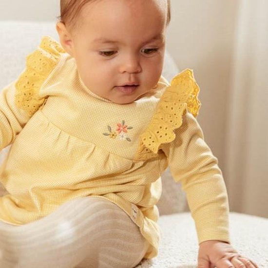 Baby met donker haar draagt een geel geruit jurkje met kanten schouderrandjes en bloemenborduring.