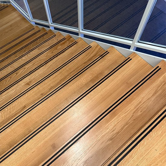 Houten trap met dubbele zwarte antislipstrips op elke trede, langs een glazen balustrade.