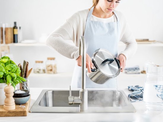 Vrouw met een lichtblauwe schort wast een roestvrijstalen kookpan met deksel af onder een kraan.