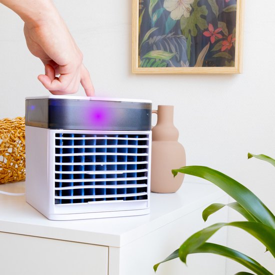 Een hand drukt op de paarse LED-verlichting van een witte mini-aircooler op een nachtkastje naast een vaas en plant.