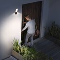 Man met tas opent deur, verlicht door een zwarte wandlamp met sensor aan de witte muur.