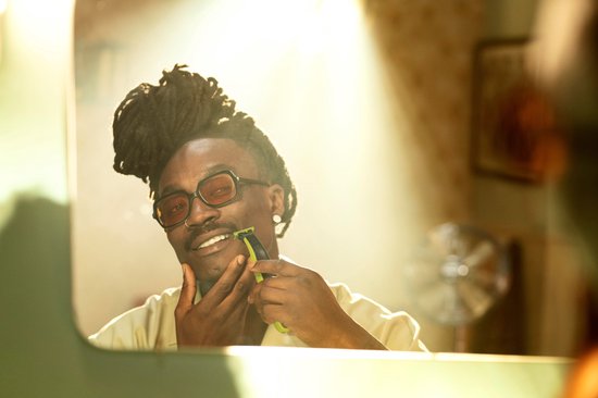Een man met dreadlocks en oranje bril trimt zijn baard met een felgroen OneBlade scheerapparaat voor een spiegel.