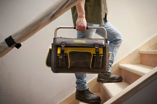 Man met spijkerbroek en werkschoenen draagt een zwarte en gele STANLEY FatMax gereedschapstas op een trap.