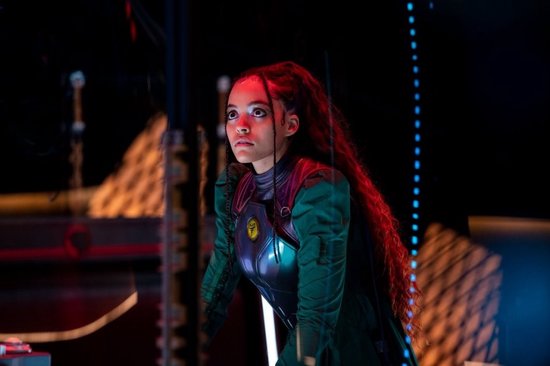 Een jonge vrouw met lang, rood krullend haar in een groen en zilveren kostuum, kijkt met intense blik naar rechts.