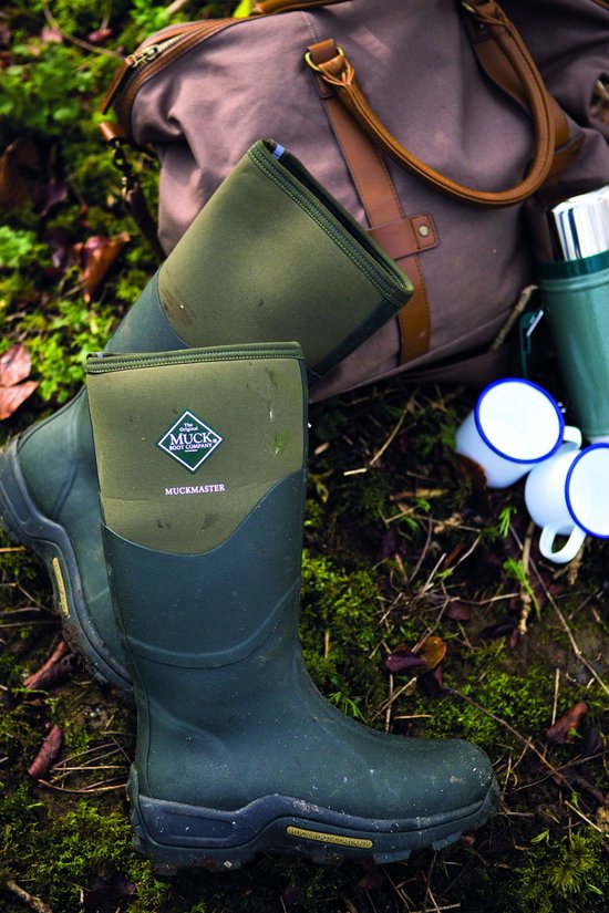 Groene Muck Boot Muckmaster laarzen liggen op mos bij een weekendtas en mokken.